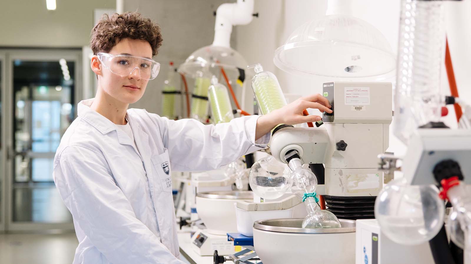 UOW medical biotechnology student Lauren Ashcroft wearing safety goggles and a white lab coat, operating a piece of laboratory equipment.