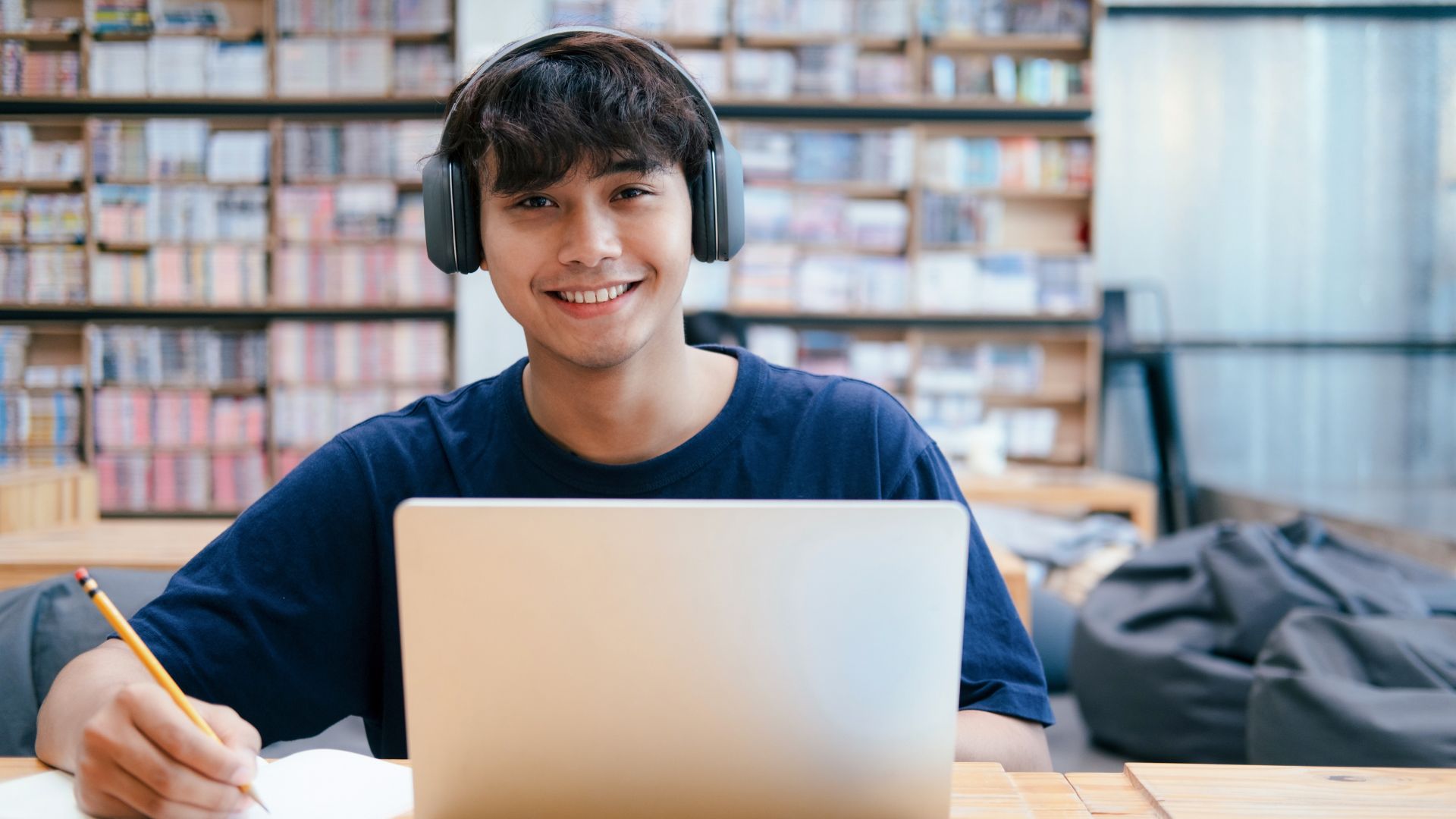 A male student sitting in a library with headphones on, a laptop in front of him, pencil in hand and smiling