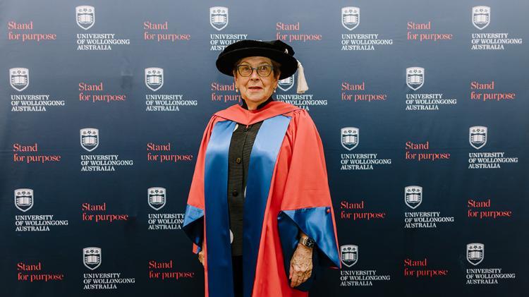 Kay Patterson in Honorary Gown in front of navy wall with repeated UOW logo