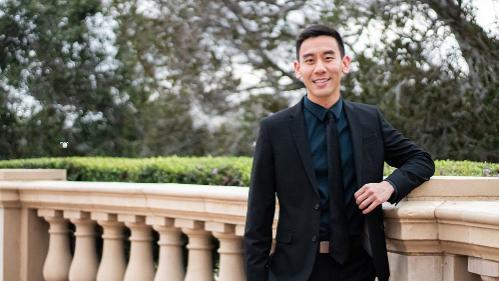 Sydney Business School, UOW Student, Bryce Lee wears a black suit and stands by a cream railing outside.