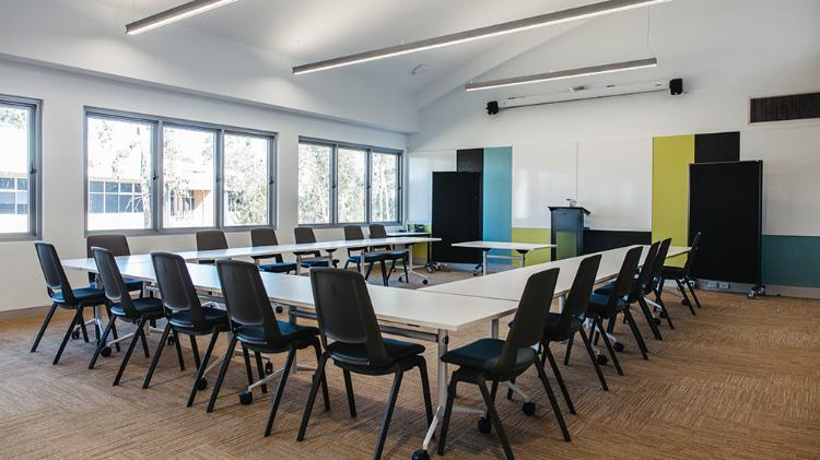 Seminar room with black chairs and white tables arranged in a horse shoe shape.