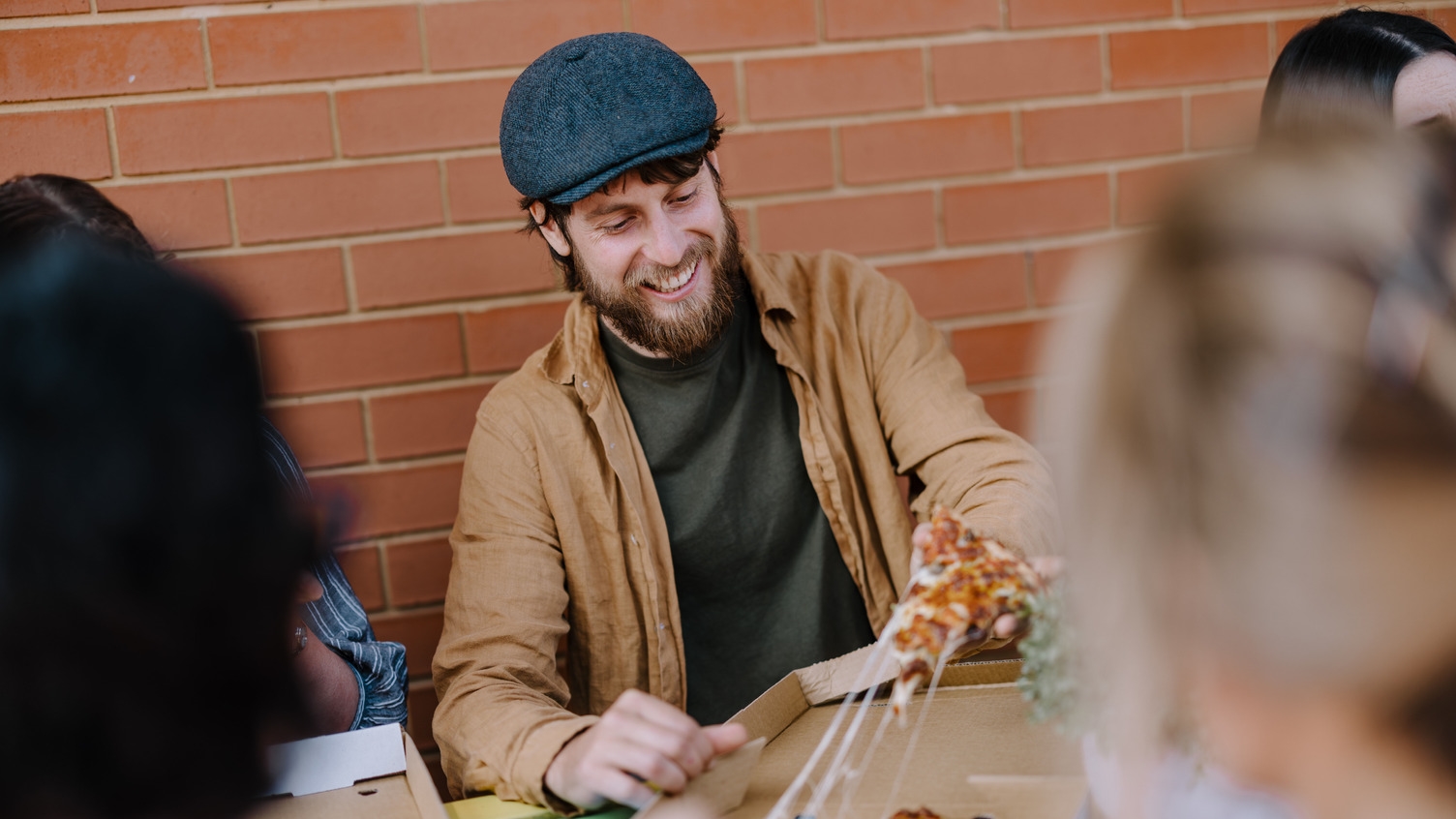 A man with a golf hat sitting at a table with a group of friends eating pizza