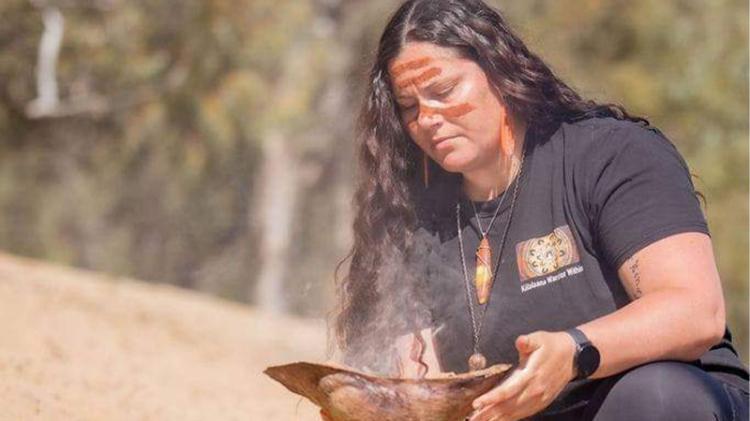 Shantelle Thompson is wearing traditional Aboriginal face paint holding a weaved basket with smoke.