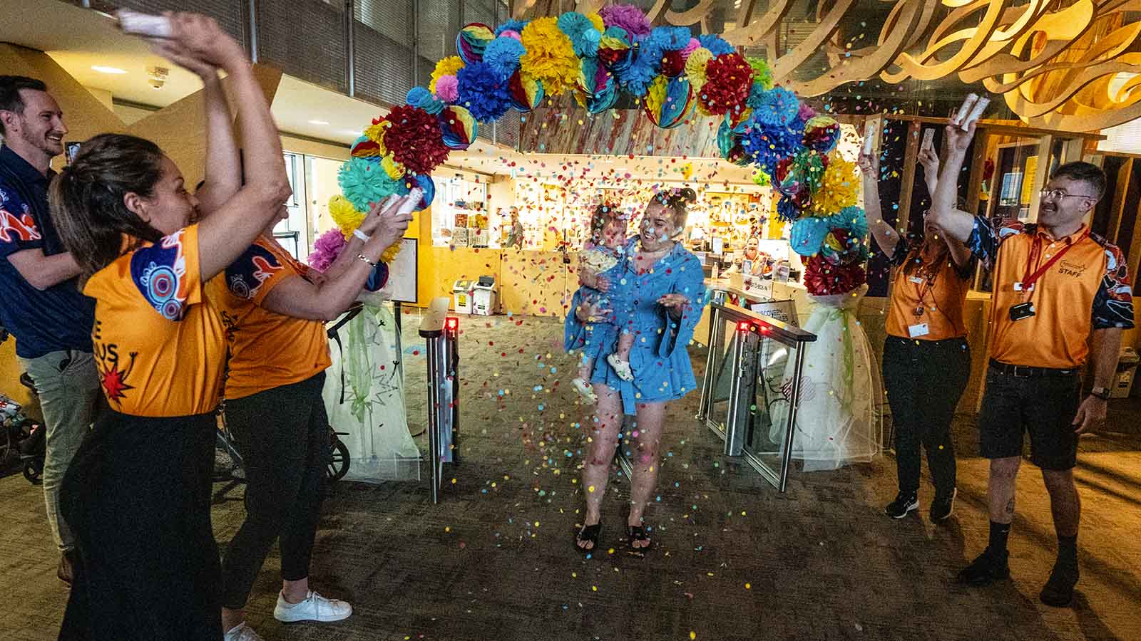 A woman and her toddler walk through an arch at Early Start Discovery Space. Onlookers throw confetti and cheer.