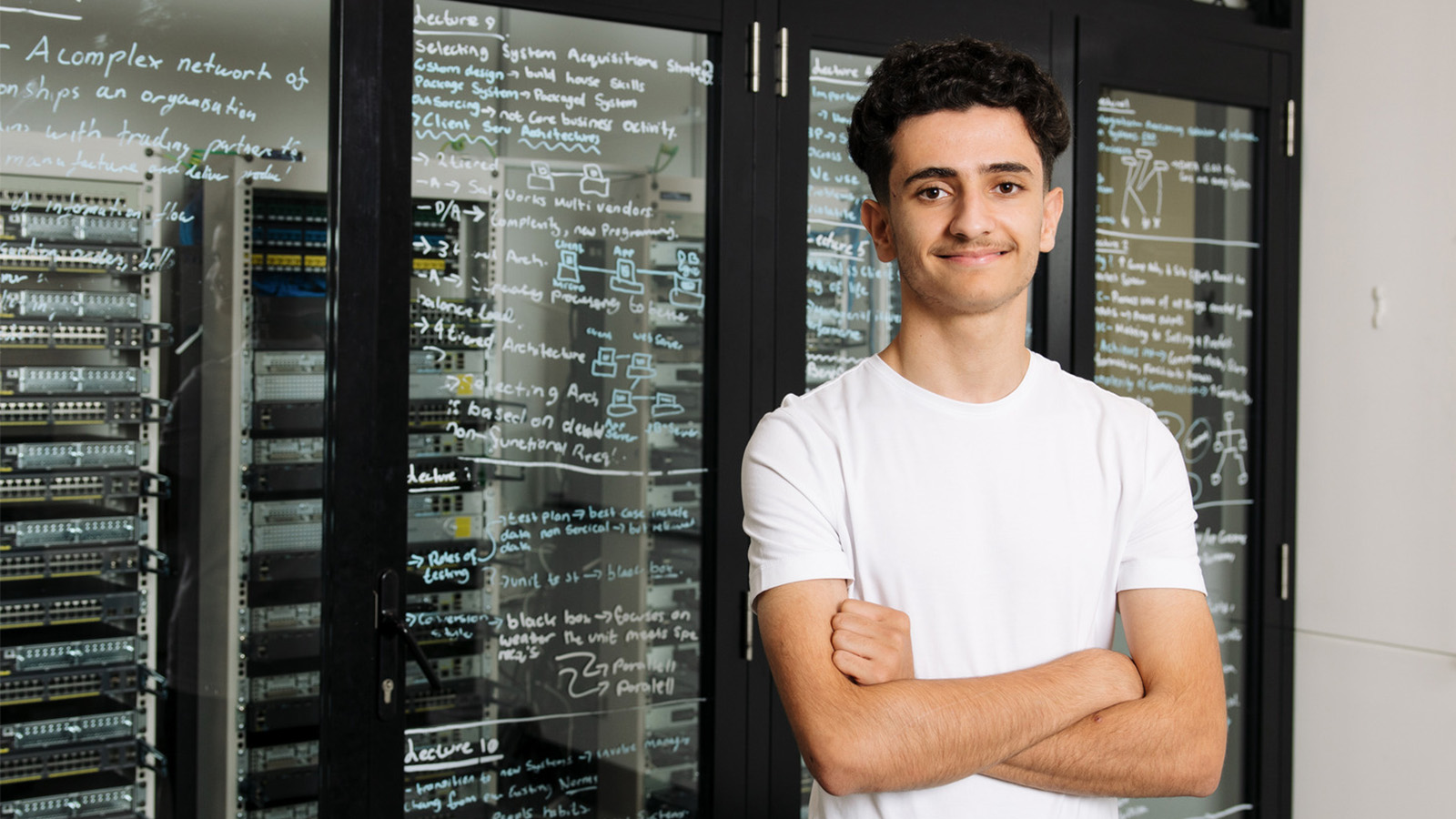 A young man in a white shirt is standing in front of a glass cabinet full of computer cables with his arms crossed.