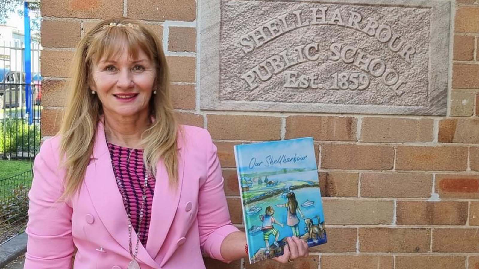 A woman is wearing a pink top and pink blazer, holding the book 'Our Shellharbour' in front from of the Shellharbour Public School sign