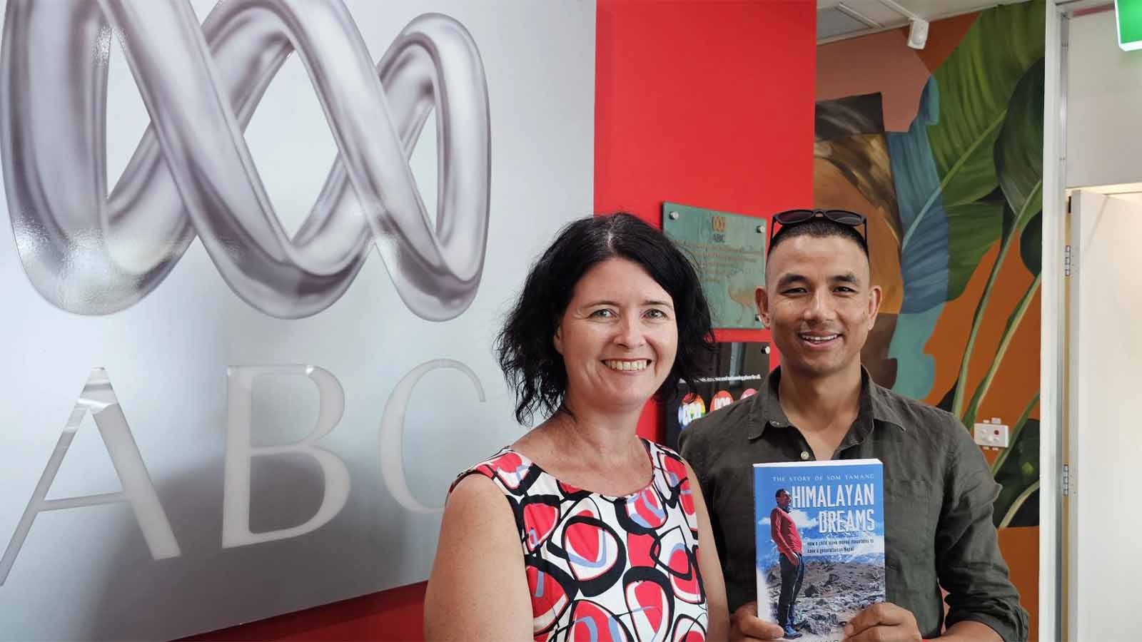 A woman and a man are standing in front of a silver ABC sign, holding the book Himalayan Dreams