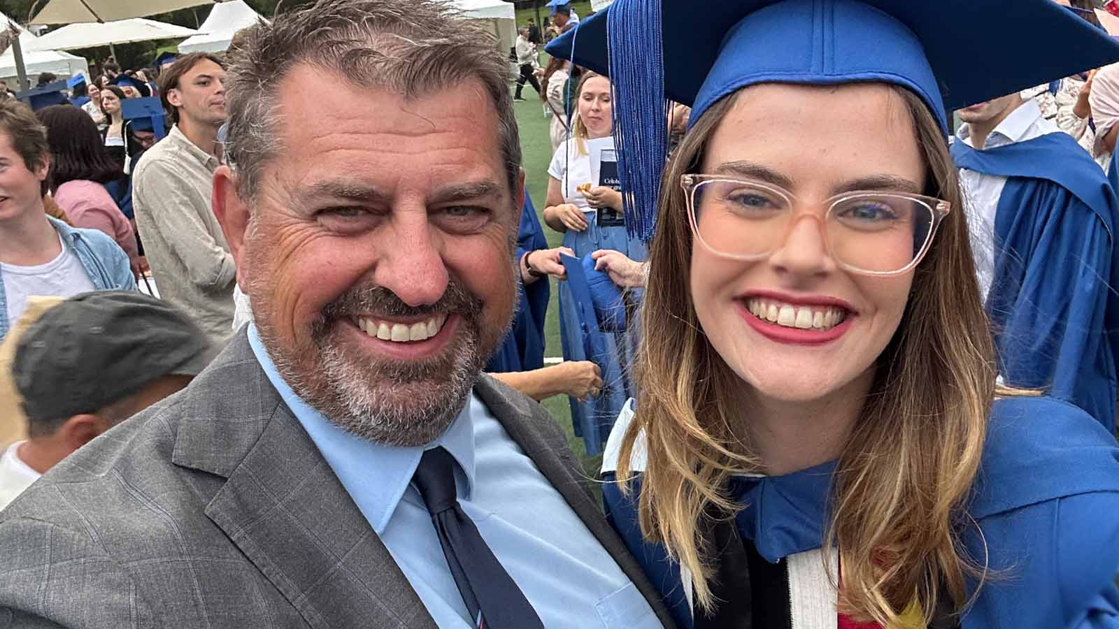 A father and daughter are smiling at the daughters university graduation. The daughter is wearing a royal blue gown and mortarbord, and black, red and yellow sash