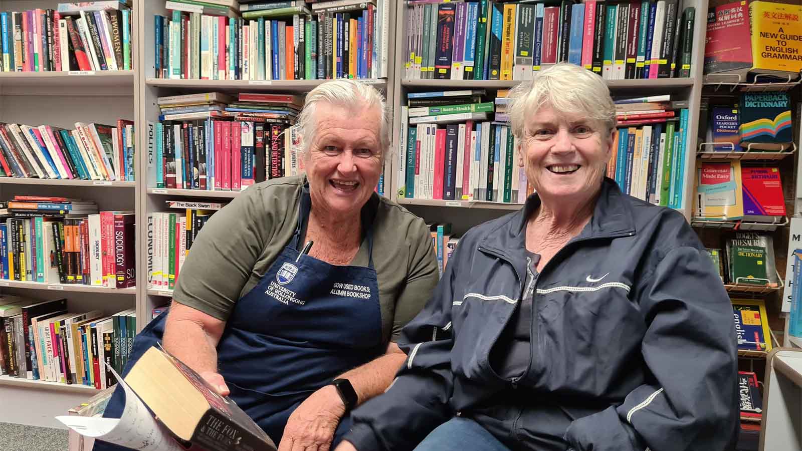 Two women are sitting in front of a shelf full of books, smiling