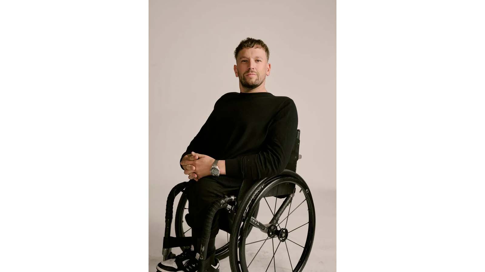 A man is sitting in a wheelchair against a cream coloured backdrop. He is wearing a black jumper and black pants with his hands clasped in his lap.