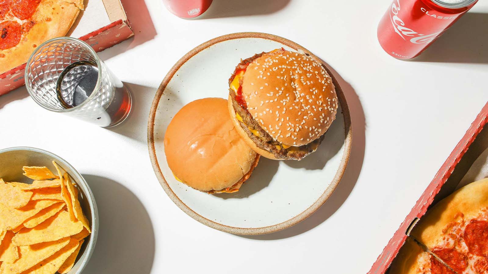Two cheese burgers are stacked on a plate on a table, surrounded by cans of Coca Cola, a bowl of corn chips and pizzas