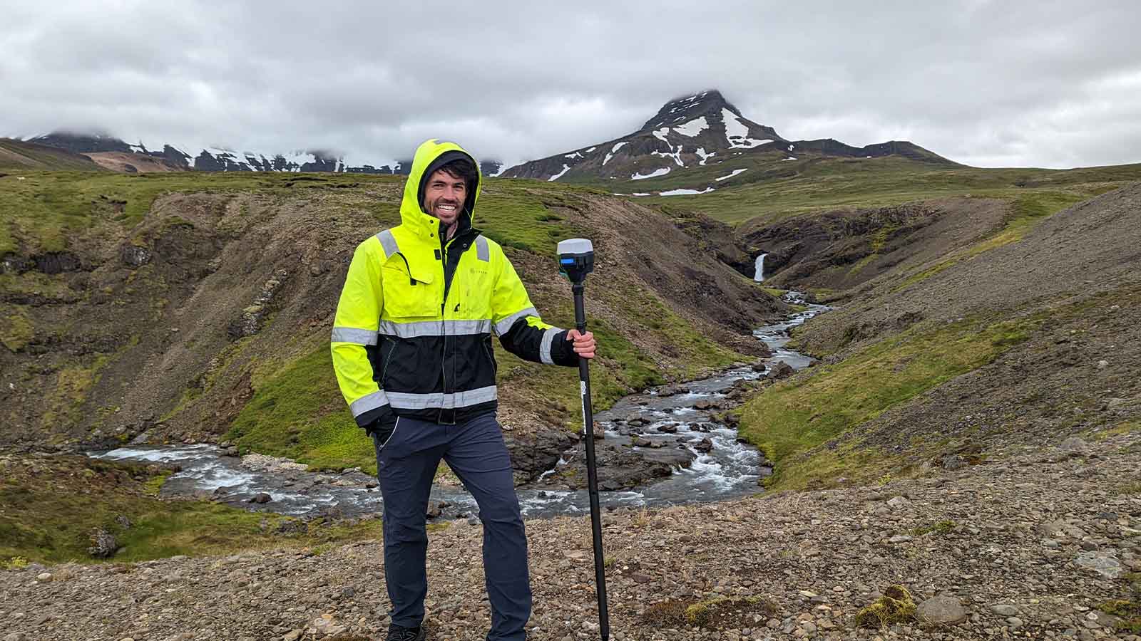 A man in a hi-vis yellow jacket is standing on a rocky landscape in Iceland, holding surveying equipment