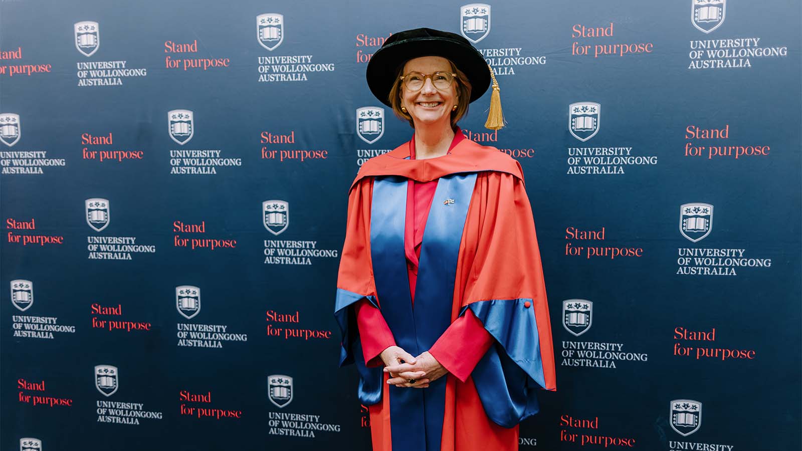 Former prime minister Julia Gillard is wearing a red and blue graduation gown and black graduation cap in front of a UOW media wall.