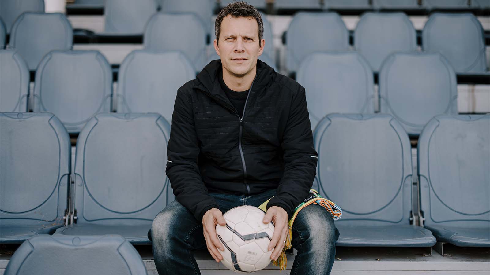 A man is sitting on stadium seating, holding a soccer ball