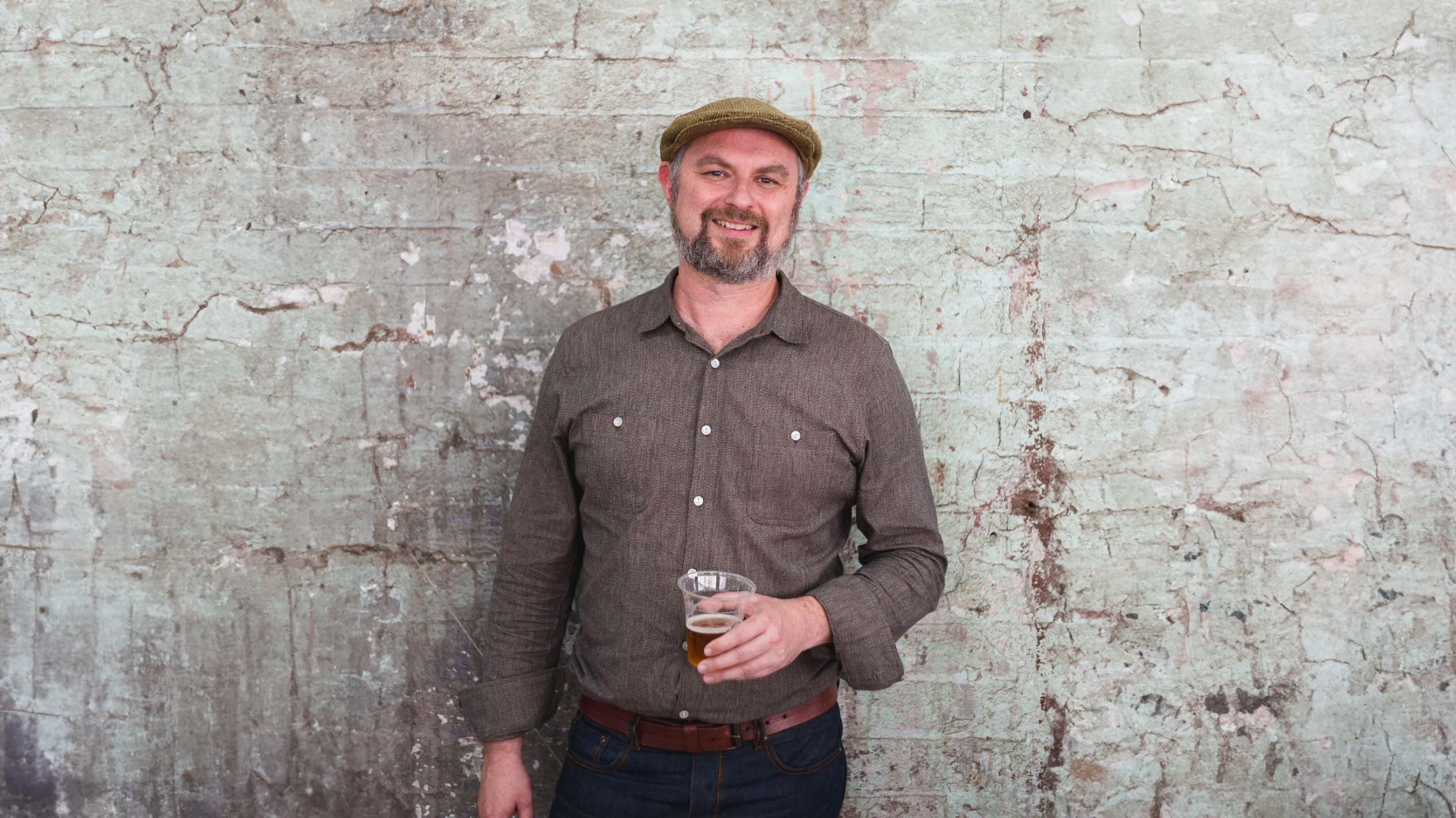 A man is standing in front of a washed out brick wall, holding a plastic cup of beer