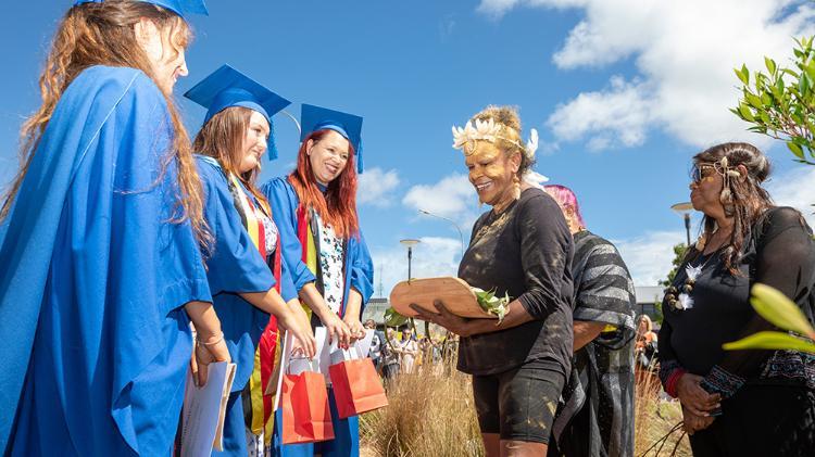 Graduates dressed in blue gowns stands outside under a a blue sky while an Aboriginal lady holds a flora wrap.