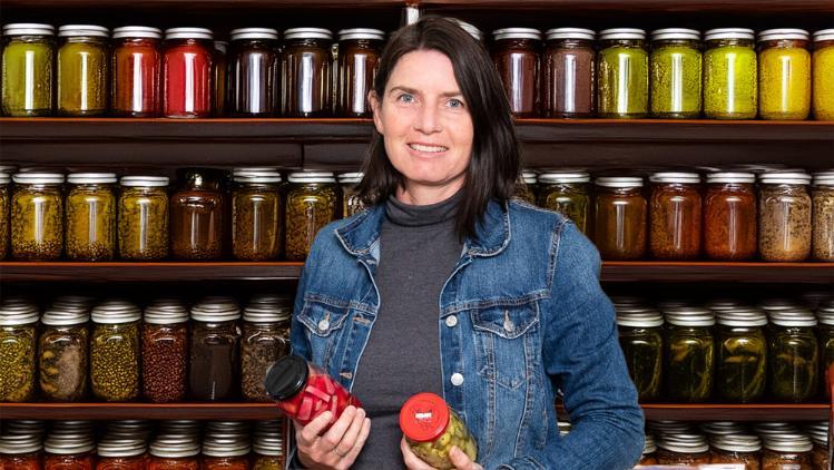 A lady is standing in front of shelves full of jars. She holds two jars in each hand.