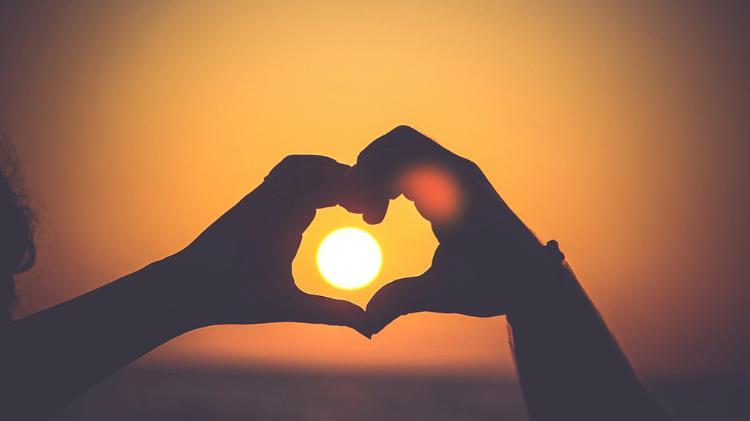 Two person making a heart space with a hand each. Orange sun is seen through the heart.