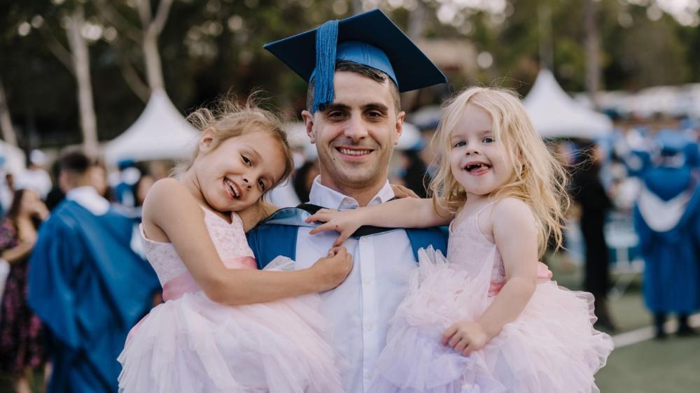 Student parent in graduation outfit holding his two daughters