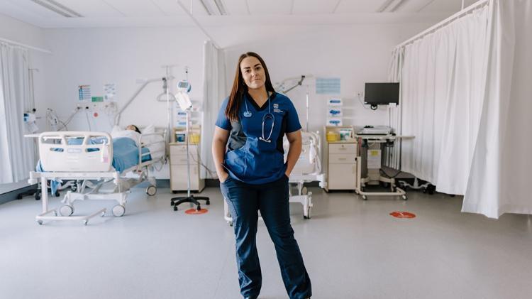 UOW Nursing student Amanda Gifford stands in a hospital room wearing her nursing uniform.