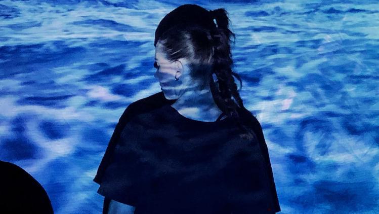 WOMan stands with her back to camera, her head slighty turned to the left. An image of water projects onto her and the wall