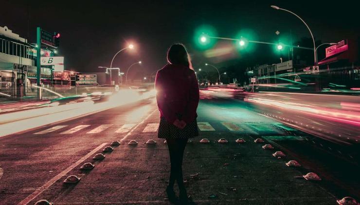 A woman stands with ther hands behind her back, in the middle of teh road as cars zoom past