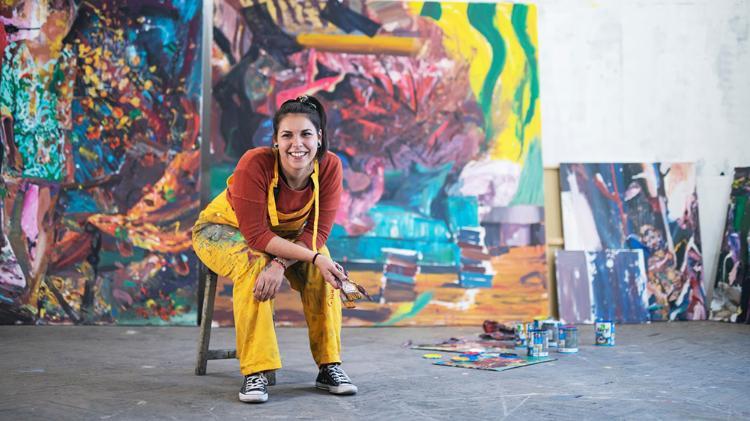A lady who is wearing yellow overalls and holding a paintbrush as she sits on a chair. A colourful swirly canvas is behind.