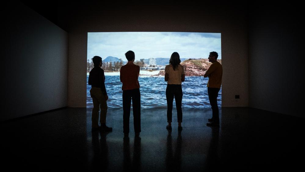 The researchers behind the City+Sea exhibition Kim Williams, Sarah Hamylton, Leah Gibbs, with Lucas Ihlein have their backs to the camera as they watch a video on the screen. . Photo: Paul Jones
