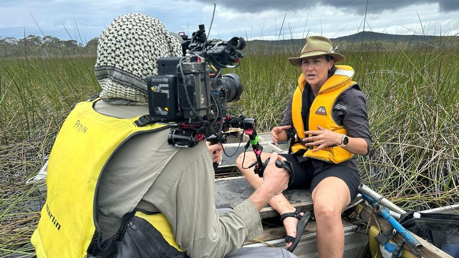 Dr Haidee Cadd sits in a boat in a wetland environment talking to the camera. In the foreground, a cameraman holds a large black camera on his shoulder. Photo: Oliver Graham