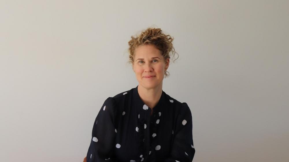 A woman wears a black and white polka dot shirt and sits against a plain wall. Photo: Jaye Murray