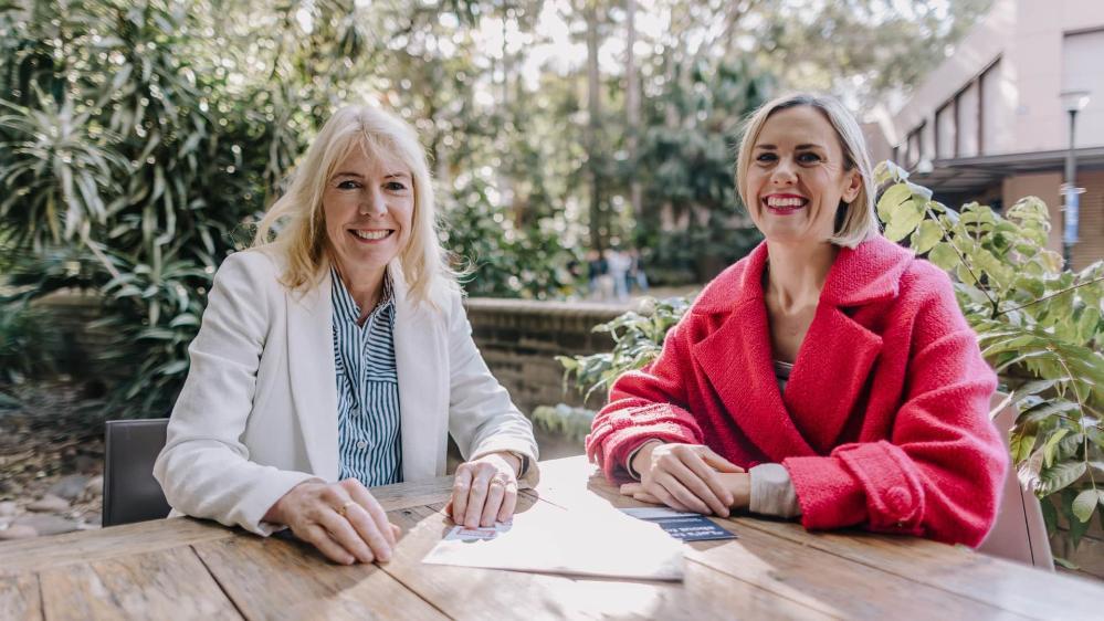 Two women sit at a cafe in an outdoor area. They are both smiling. There are pamphlets on the table in front of them. Photo: Michael Gray