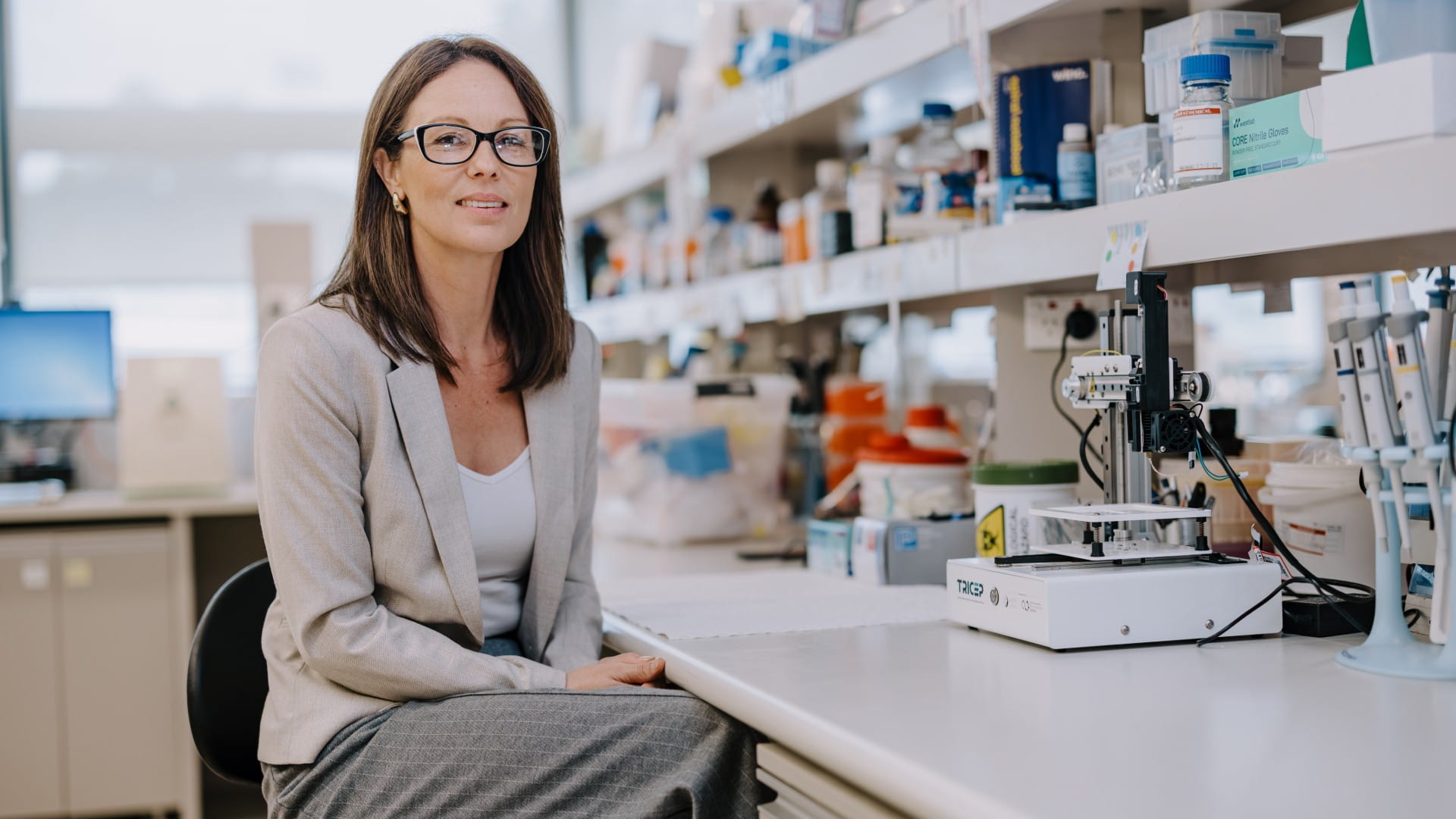 A woman wearing glasses sits at a lab bench surrounded by scientific equipment. Photo: Michael Gray