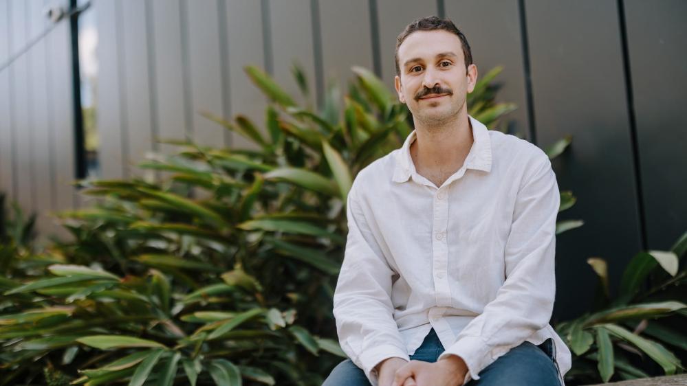 A man sits on a bench against a dark backdrop with bushes. Photo: Michael Gray