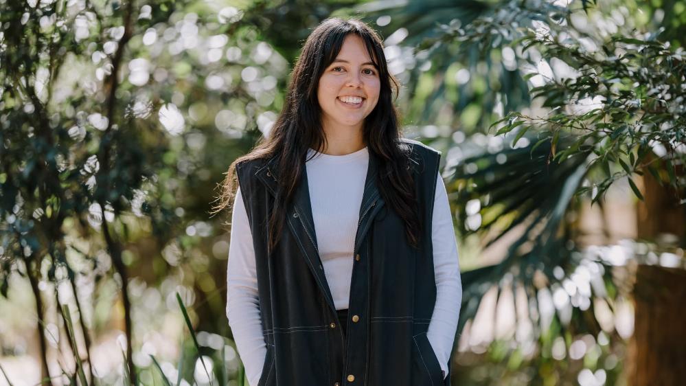 PhD candidate Naomi May stands and smiles, against a backdrop of bushes. Photo: Michael Gray