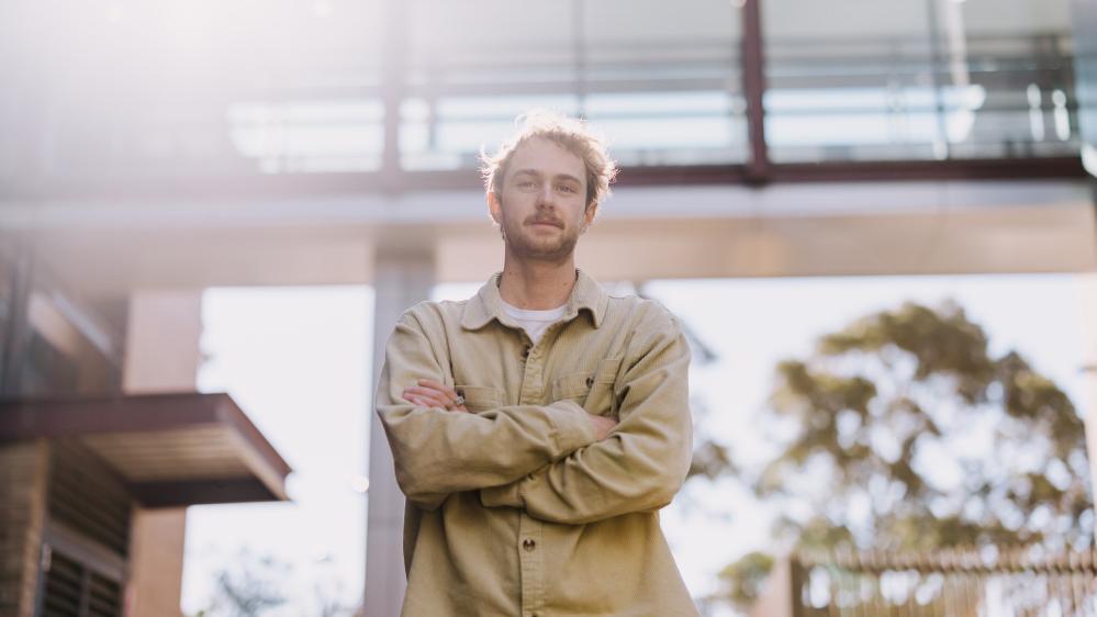 A man stands in front of a building, with the sun in the background, wearing a beige jacket. His arms are crossed. Photo: Michael Gray