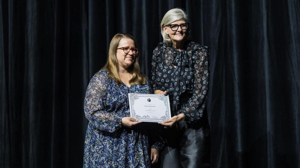 A woman stands on a stage and receives a certificate from another woman. They are both smiling. Photo: Ben Calvert Photography